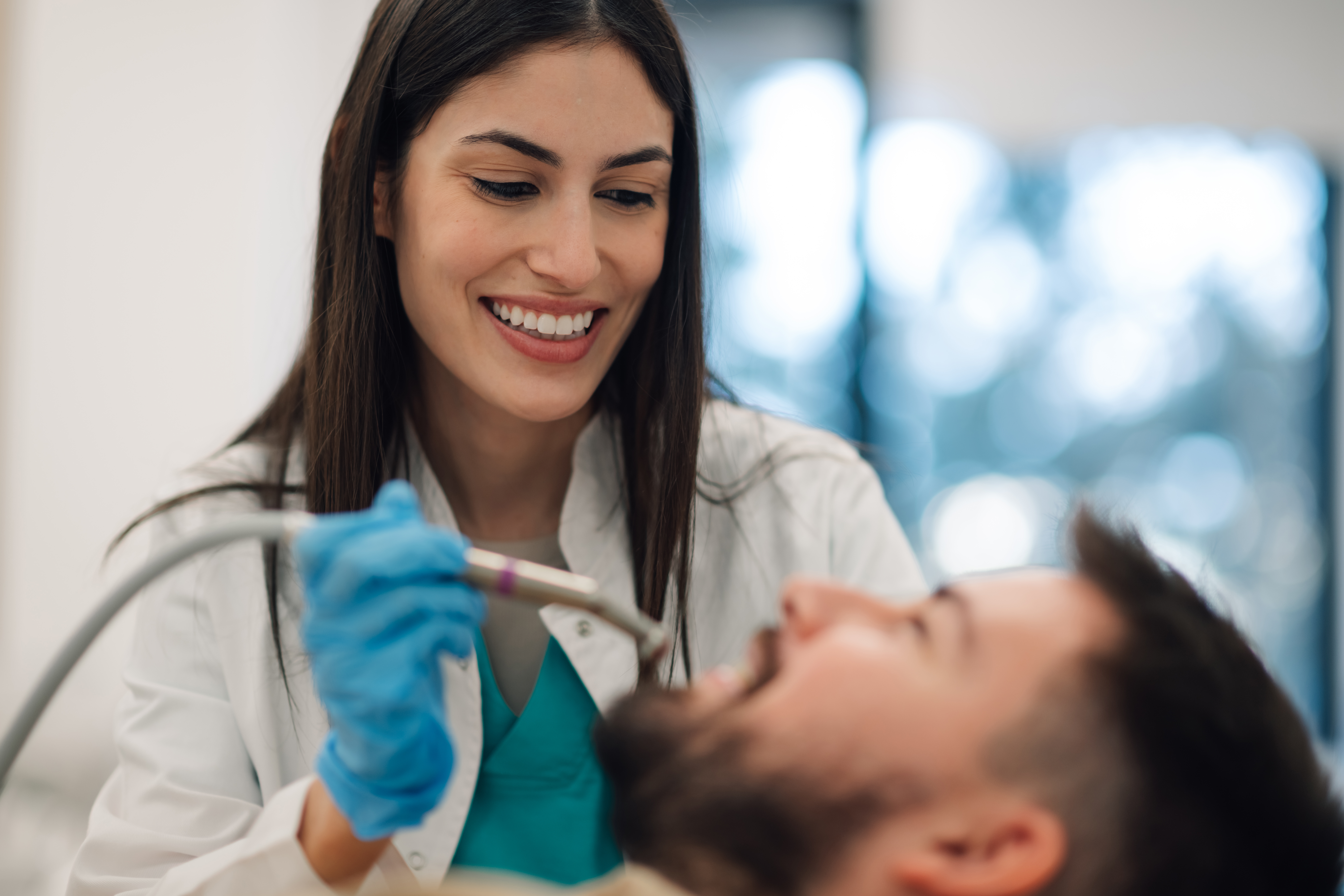 Smiling female dentist wearing gloves and holding a dental drill, performing a dental procedure on a male patient in a modern dental clinic, ensuring optimal oral health and hygiene
